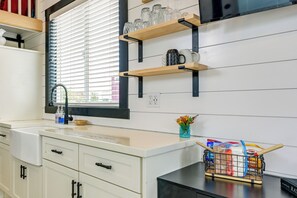 A kitchen with white cabinets, a black countertop, and a window with blinds. There are shelves above the counter holding glassware and a small vase with flowers.