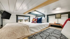 Un dormitorio con cama, mesita de noche, lámpara, televisor y tragaluz.