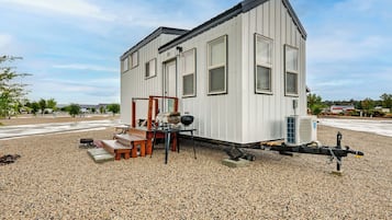 Ein eingeschossiges Gebäude mit flachem Dach, einer kleinen Veranda mit Sitzbank und einem tragbaren Grill auf Kiesgrund.