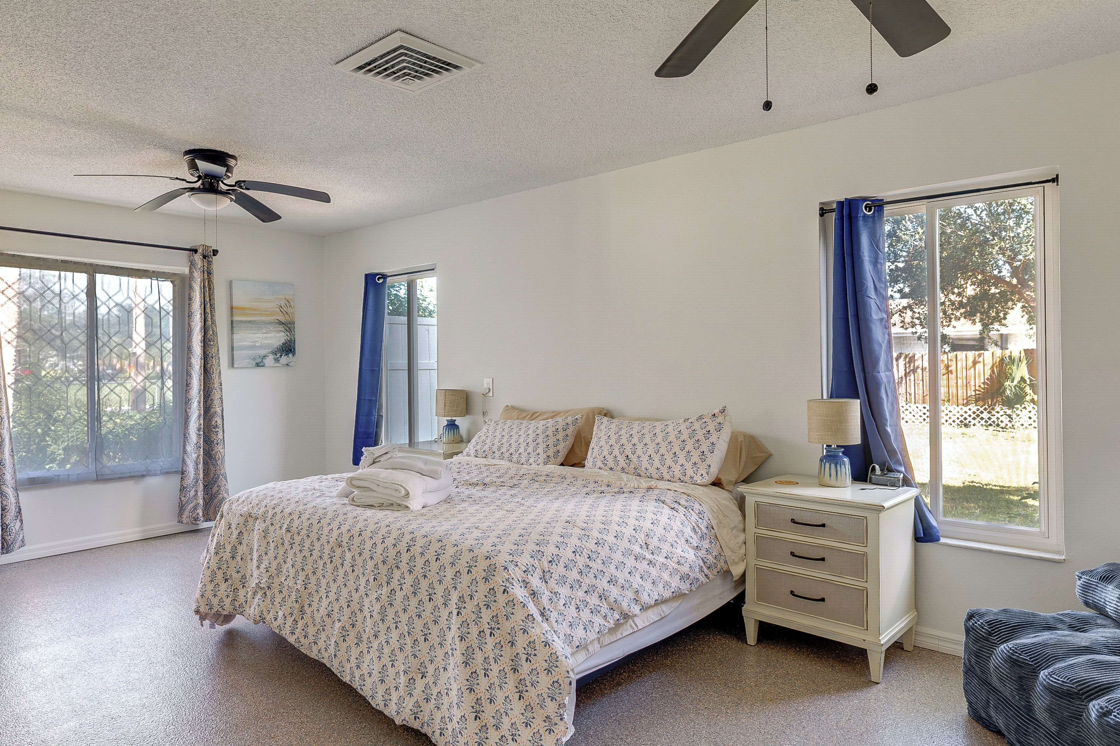 A bedroom with a bed, nightstand, lamp, ceiling fan, and large windows.