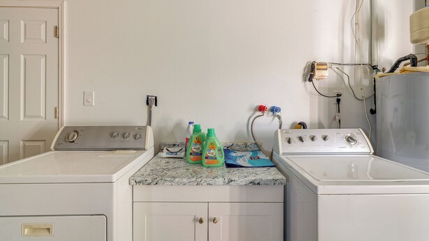 Uma lavanderia com duas máquinas de lavar, um aquecedor de água e diversos produtos de limpeza no balcão.