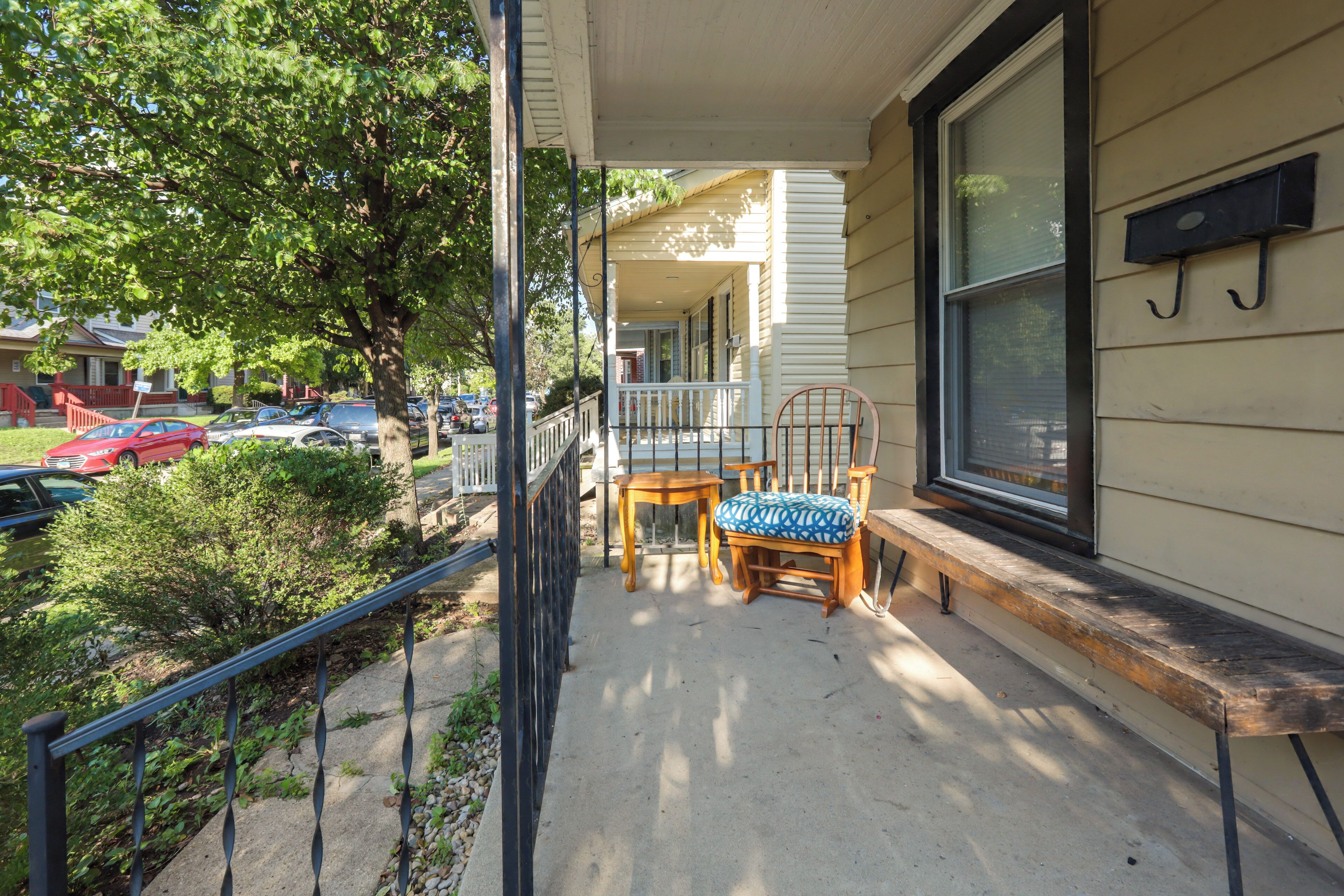 A covered porch with a table and chairs, a window with a hanging sign, and a view of parked cars and houses.