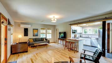 A modern living room with a sofa, coffee table, and a TV. There is a kitchen area with a dining table and bar stools.