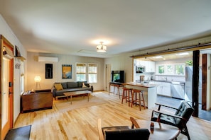 A modern living room with a sofa, coffee table, and a TV. There is a kitchen area with a dining table and bar stools.