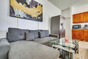 A living room with a grey sofa, a glass coffee table, and a kitchen area with wooden cabinets and a stainless steel refrigerator.