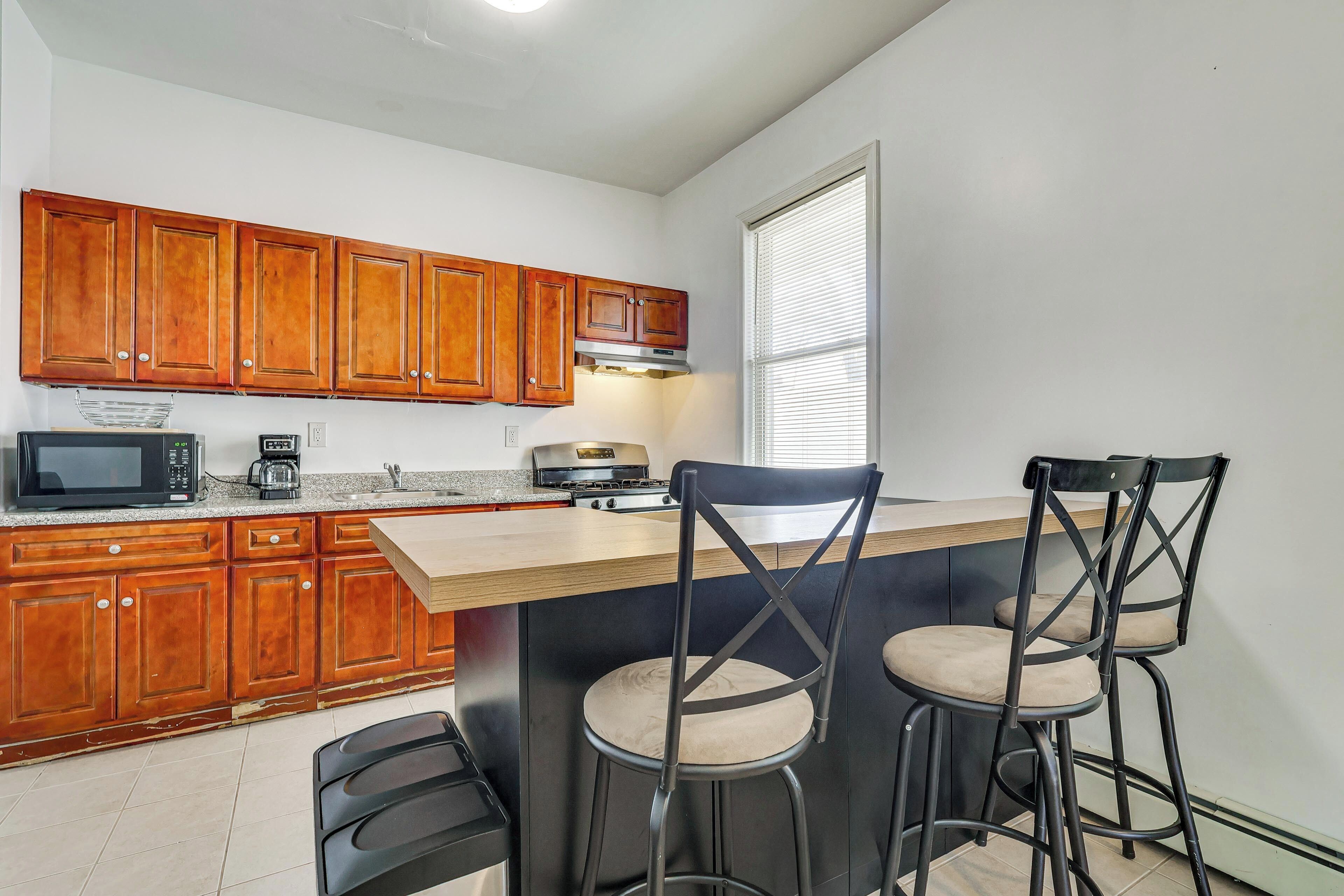 A kitchen with wooden cabinets, a microwave, a coffee maker, and a stove. There are three bar stools with cushioned seats.