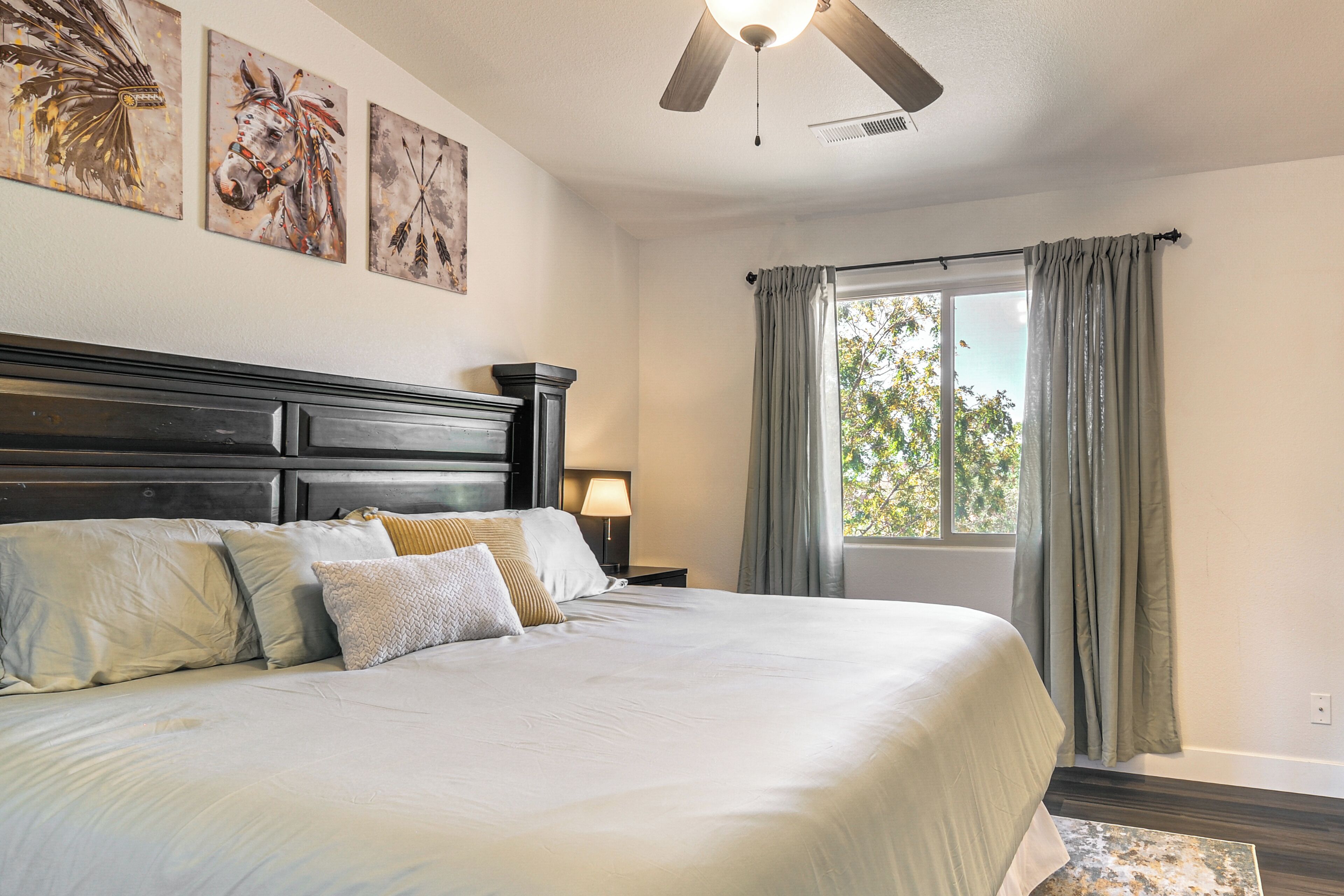 A bedroom with a large bed, a ceiling fan, a window with curtains, and artwork on the wall.