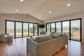 A spacious living room with a large grey sofa, a television mounted on the wall, and a view of the ocean through floor-to-ceiling windows.