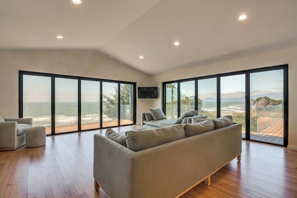 A spacious living room with a large grey sofa, a television mounted on the wall, and a view of the ocean through floor-to-ceiling windows.