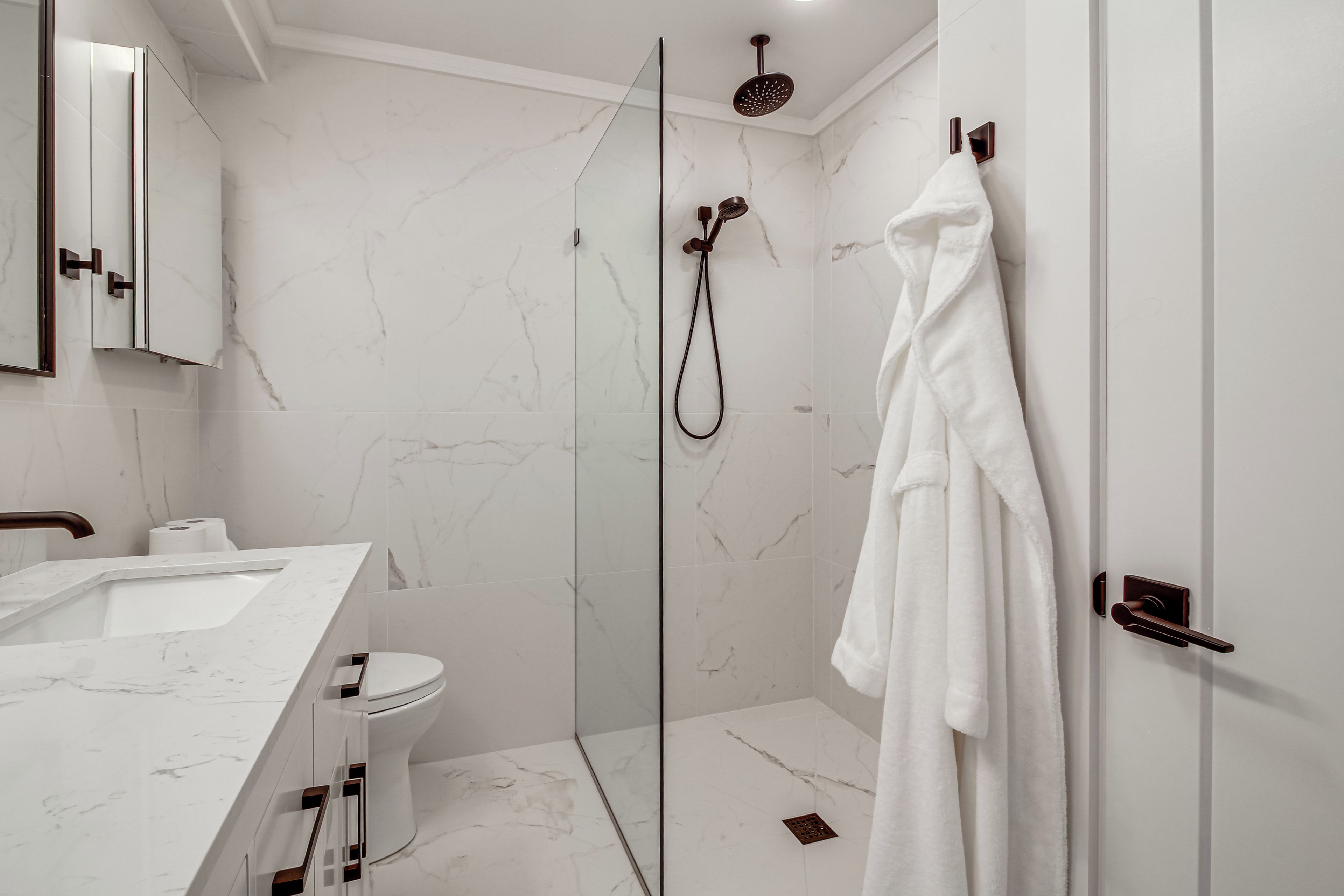 A modern bathroom with a glass-enclosed shower, a white marble sink, and a white bathrobe hanging on the door.