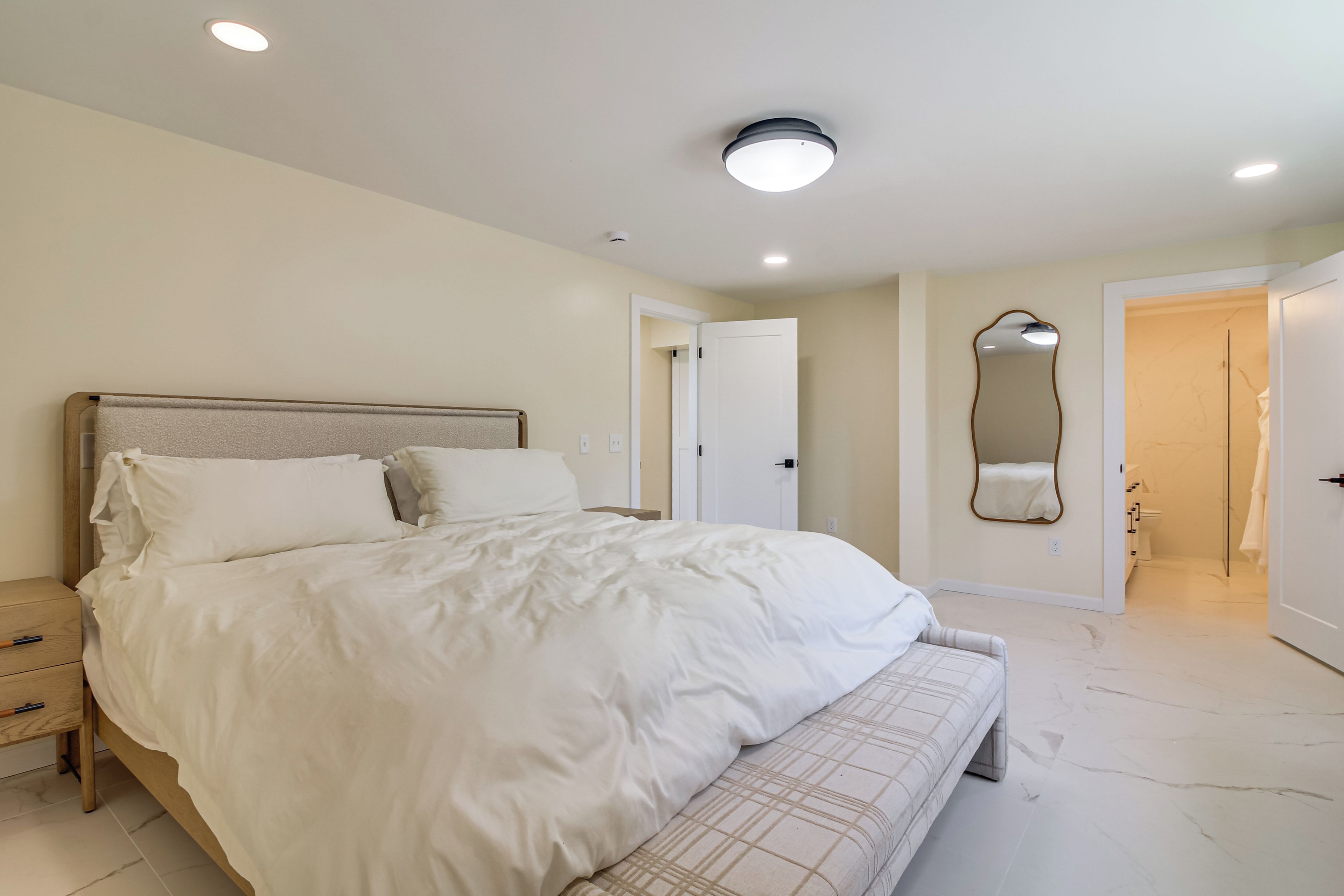 A bedroom with a large bed, bedside tables, a bench, a mirror, and a door leading to a bathroom.