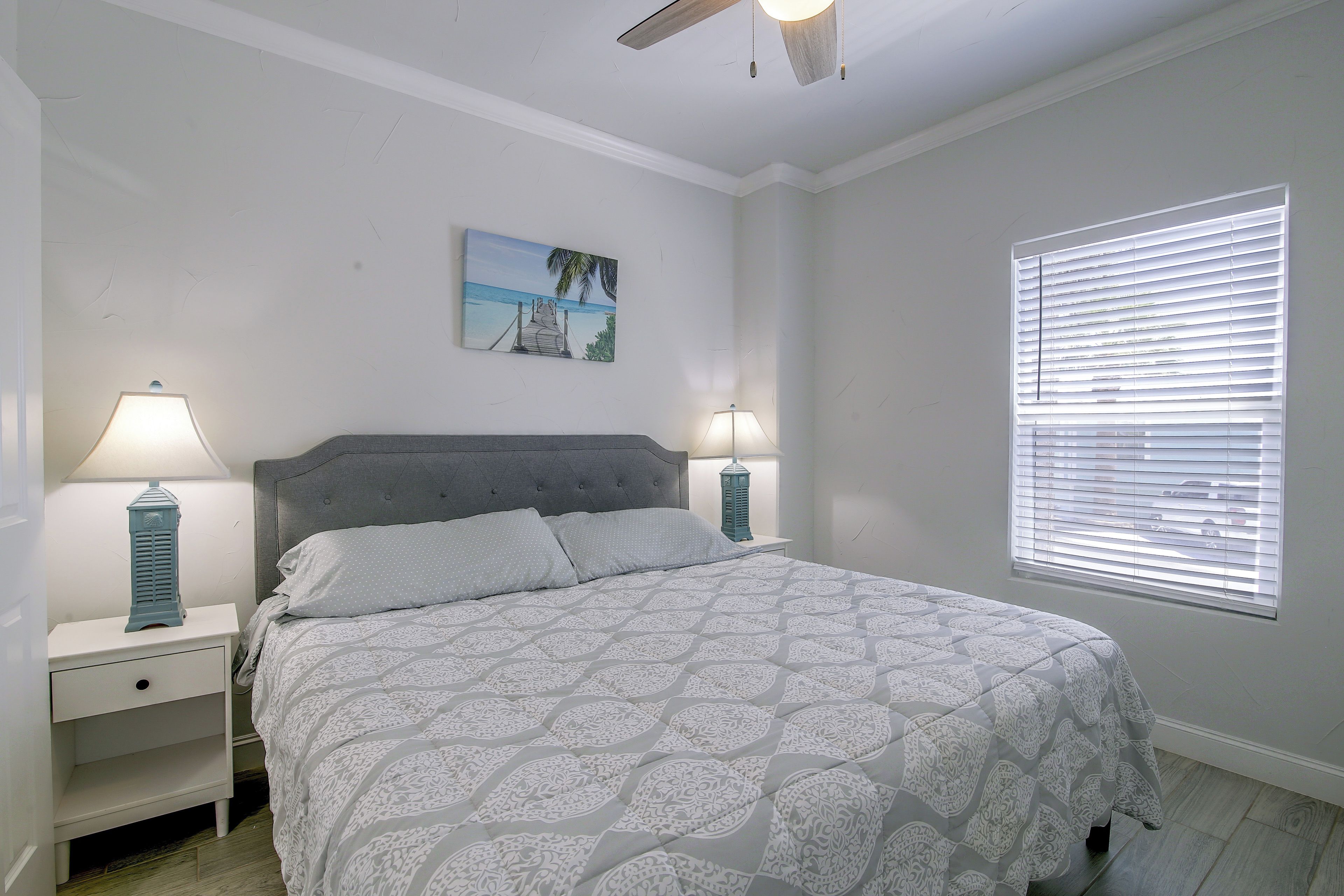 A bedroom with a bed, two bedside tables, a nightstand, a ceiling fan, a window with blinds, and a wall-mounted picture.