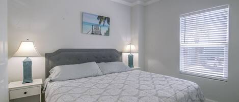 A bedroom with a bed, two bedside tables, a nightstand, a ceiling fan, a window with blinds, and a wall-mounted picture.