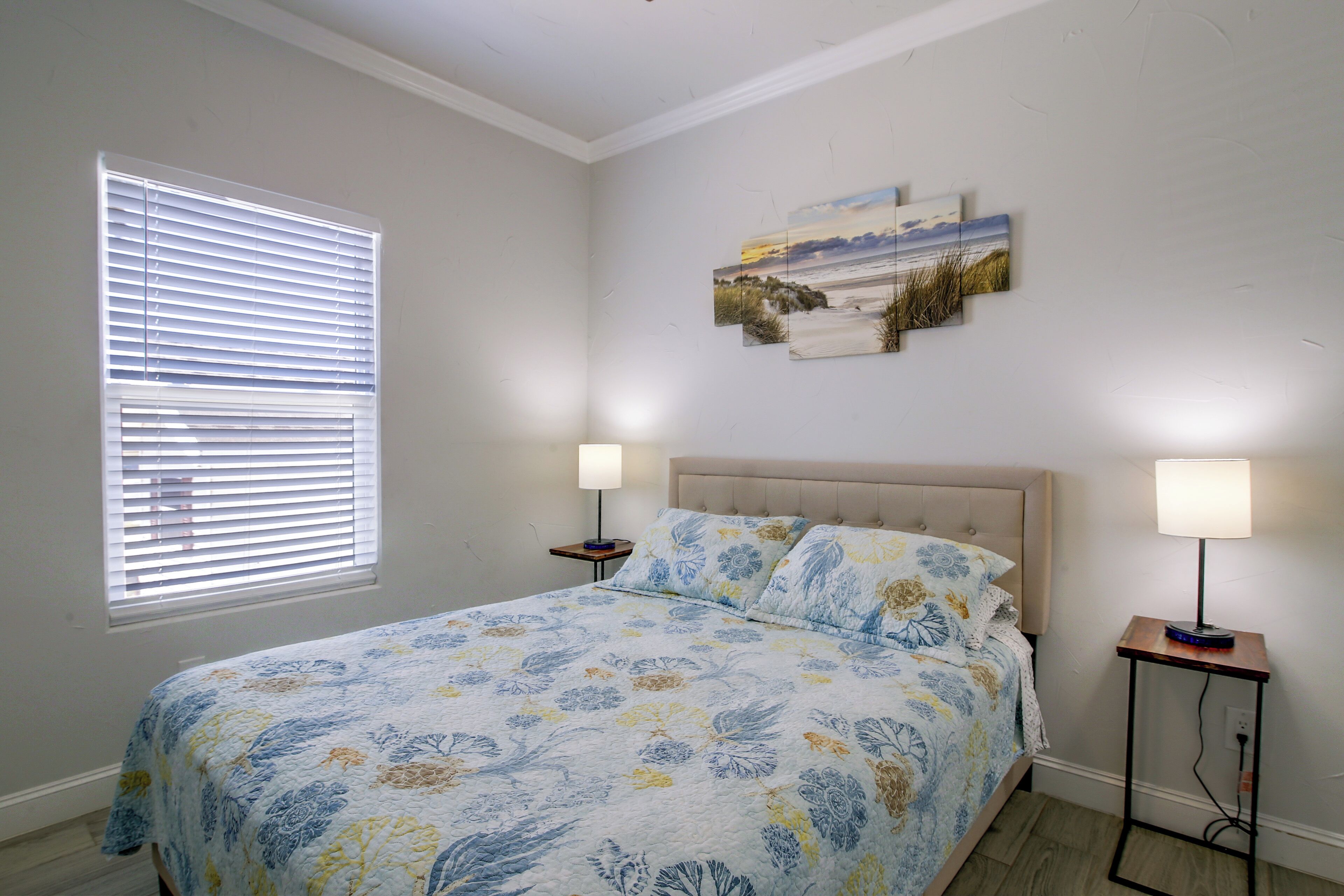 A bedroom with a bed, two bedside lamps, a window with blinds, and a wall-mounted artwork.