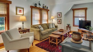 A living room with a sofa, armchair, coffee table, and a television. There are two lamps, a ceiling fan, and a large window with blinds. - Deck & Wooded Views! 2 Mi to Frederick Gunn School (Washington)
