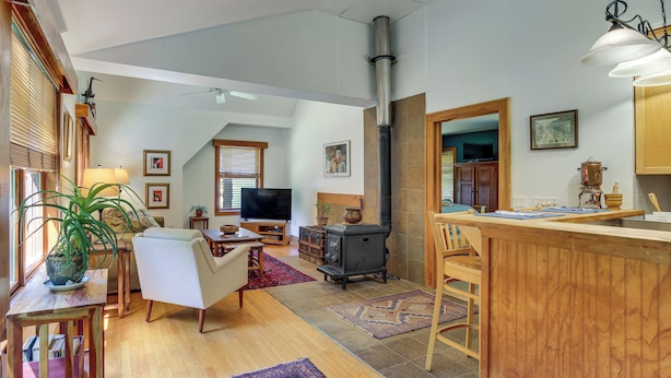 A spacious living area with a wooden floor, a television, a sofa, a dining table, and a kitchen area with a stove. - Deck & Wooded Views! 2 Mi to Frederick Gunn School (Washington)