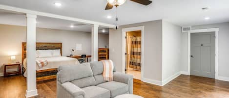 Une pièce spacieuse avec un ventilateur de plafond, un lit doté d’une tête de lit en bois, un canapé gris, une petite table ronde et une salle de bain visible à travers une porte ouverte.