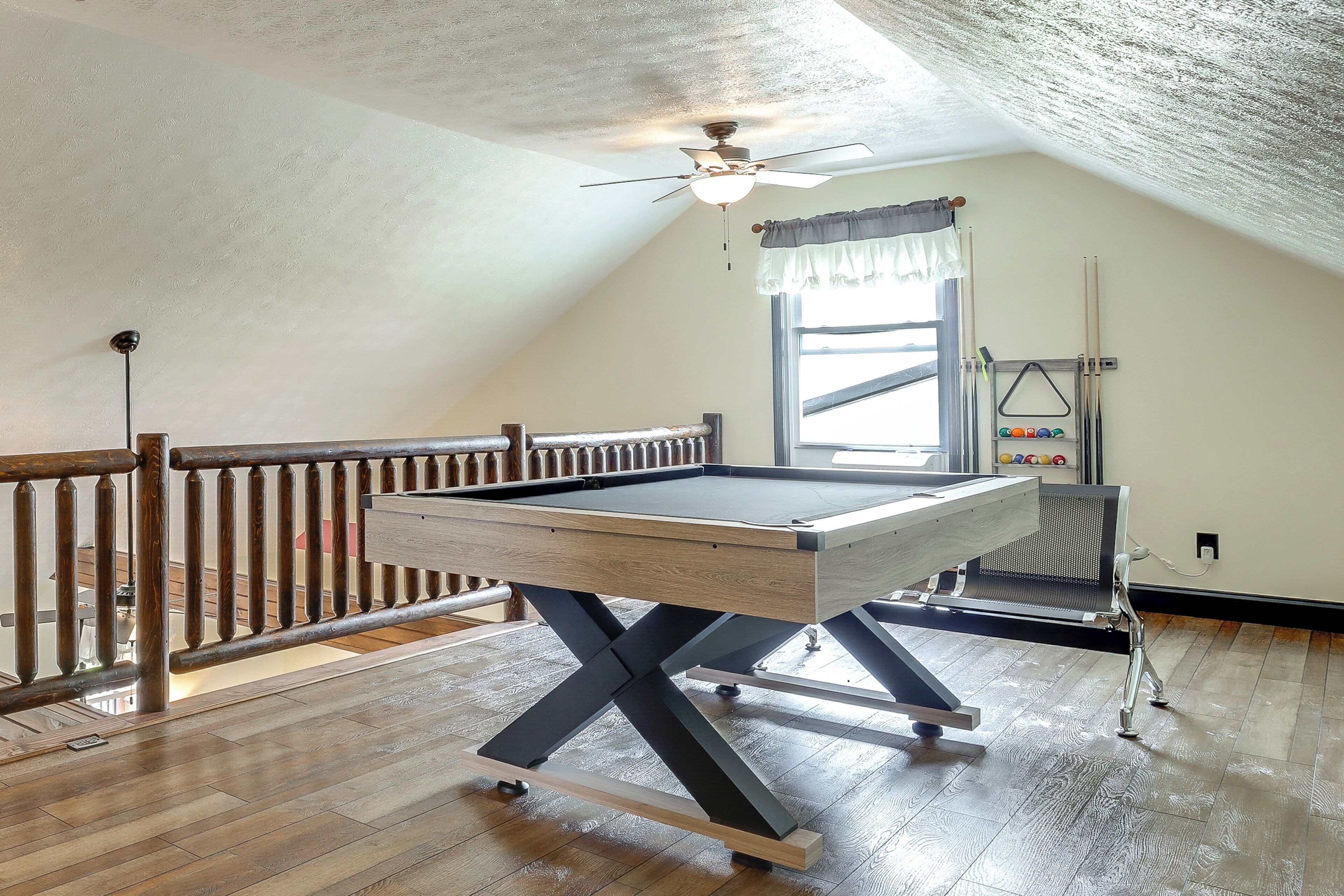 A pool table in a room with wooden flooring, a ceiling fan, and a window with curtains.