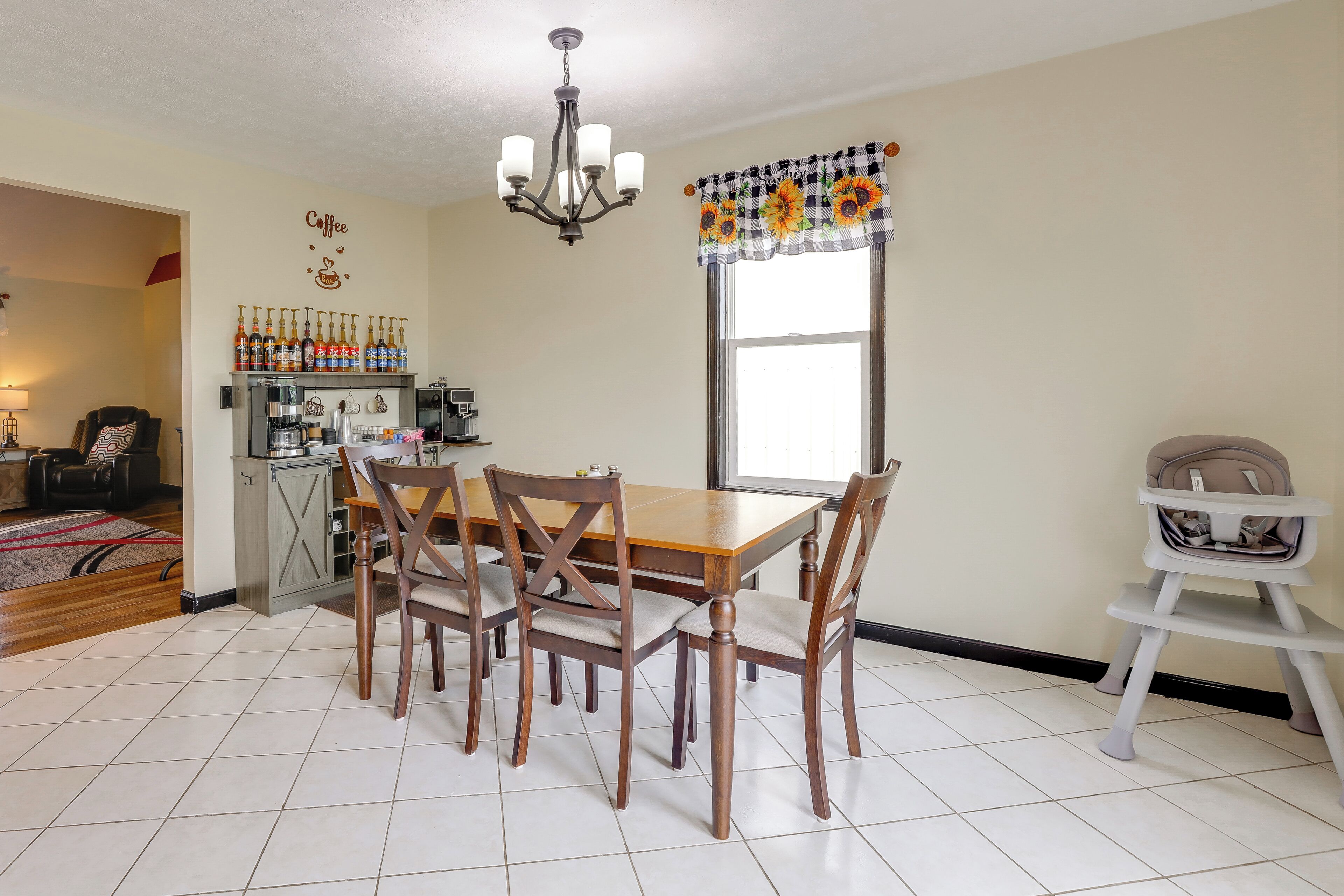 A dining area with a wooden table and chairs, a chandelier, a kitchen area with coffee-making equipment, and a window with a sunflower curtain.