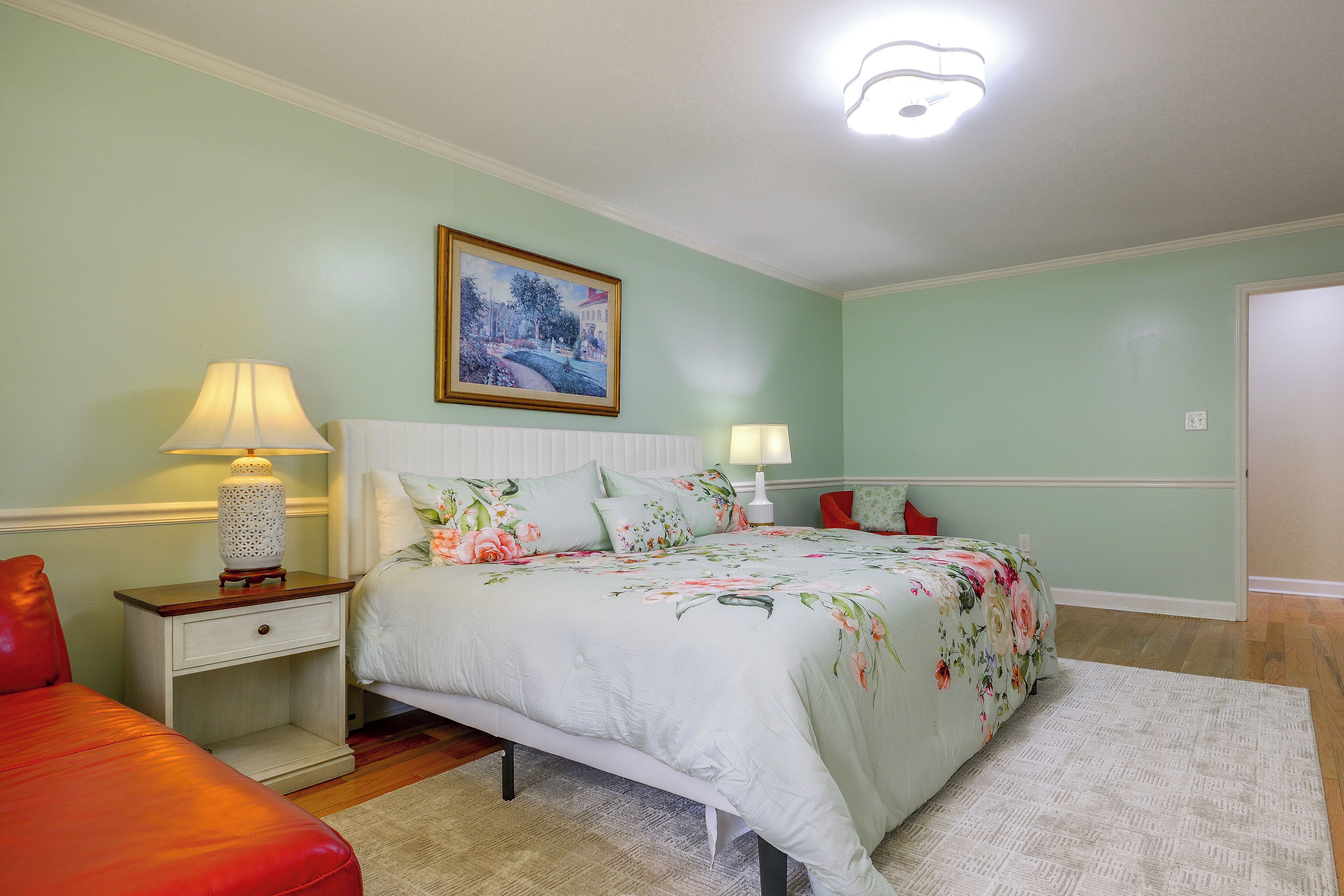A bedroom with a bed, nightstands, a red chair, a painting on the wall, and a door.