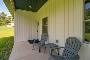 A covered porch with two Adirondack chairs, a door, and a window overlooking a lawn.