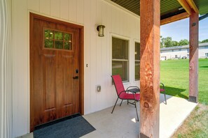 A wooden front door with a glass panel, a red chair, and a small table on a porch with a white exterior wall and a green lawn visible in the background.