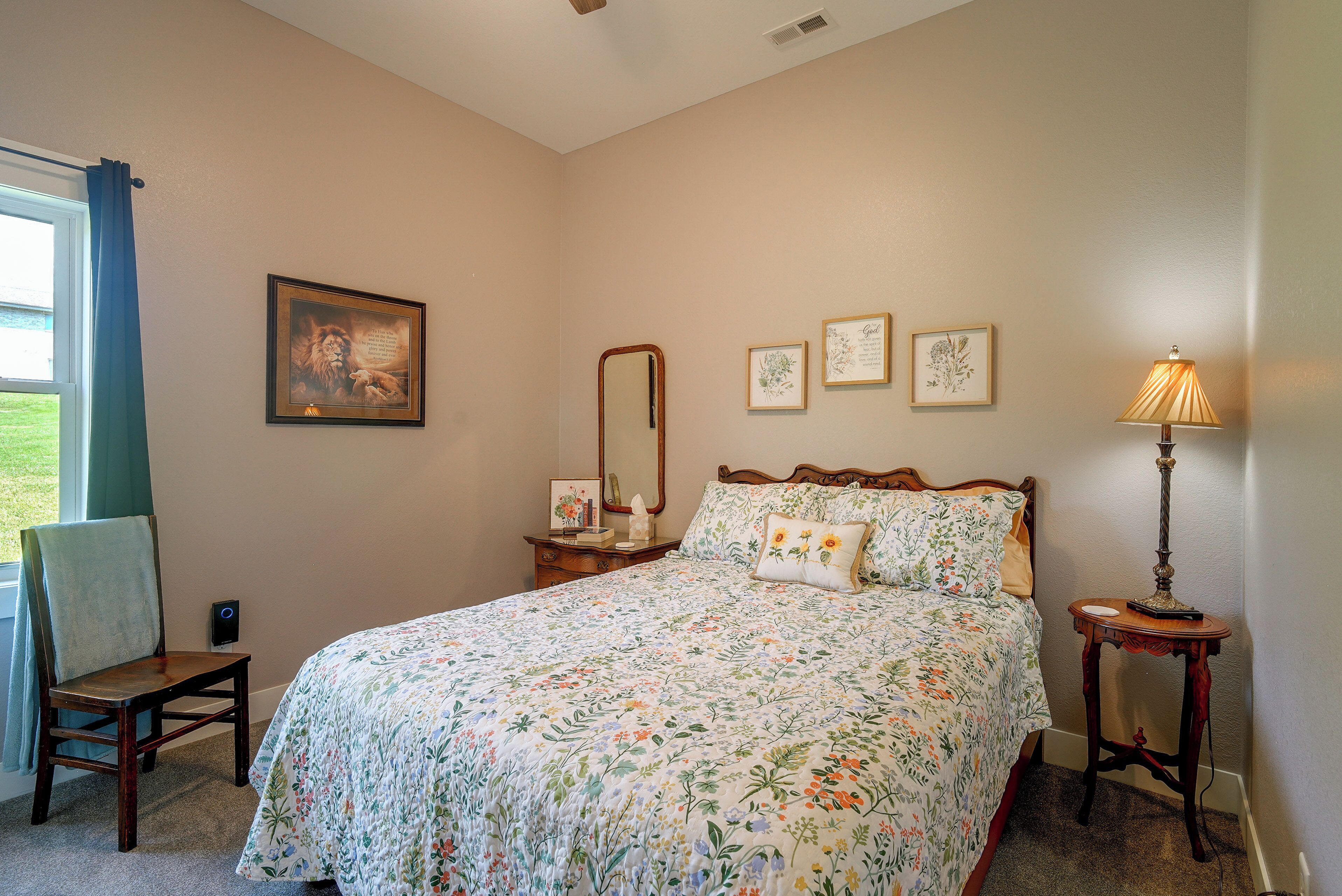 Un dormitorio con colcha estampada, una mesita de noche con lámpara, una silla, una ventana con cortinas y decoración en la pared.