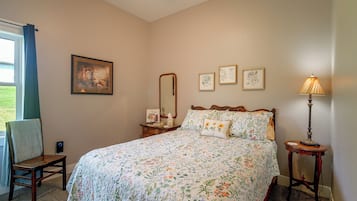 Un dormitorio con colcha estampada, una mesita de noche con lámpara, una silla, una ventana con cortinas y decoración en la pared.