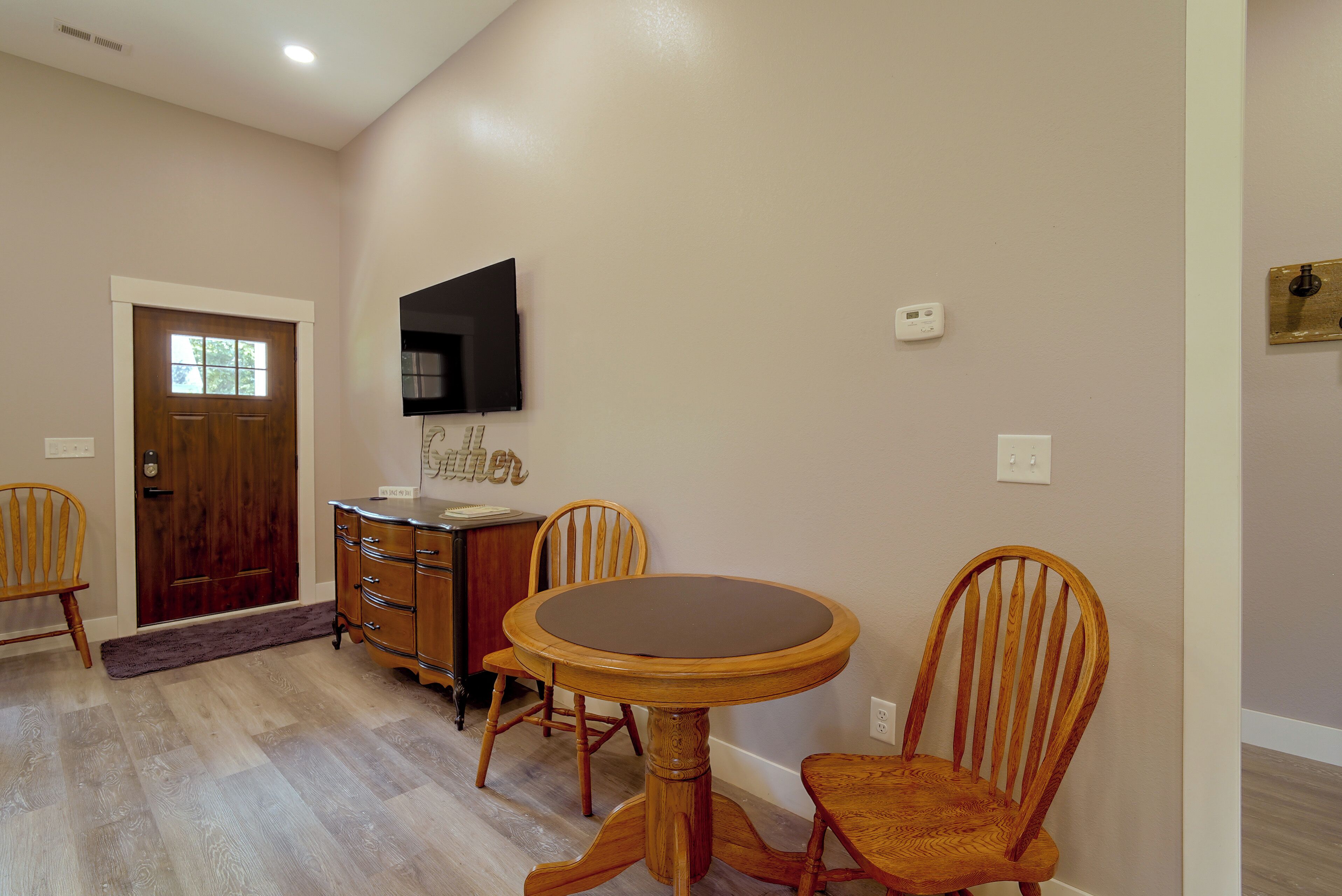 A dining area with a round table and four chairs, a wooden cabinet, a flat-screen TV mounted on the wall, and a door with a window.