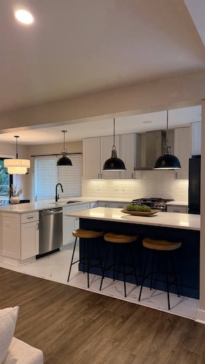 A modern kitchen with a central island, bar stools, pendant lighting, and a dishwasher.