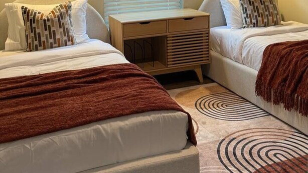 A bedroom with two beds, a ceiling fan, bedside lamps, a window with blinds, a bedside table, and a patterned rug.