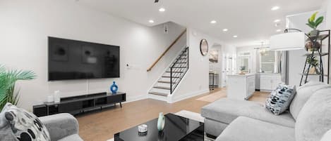 Un salon moderne avec un téléviseur à écran plat, une table basse noire, des canapés gris, un escalier et une cuisine en arrière-plan.