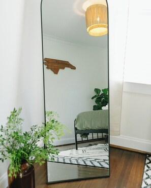 Quarto com espelho grande, cama com colcha verde, prateleira de madeira e luminária pendente.