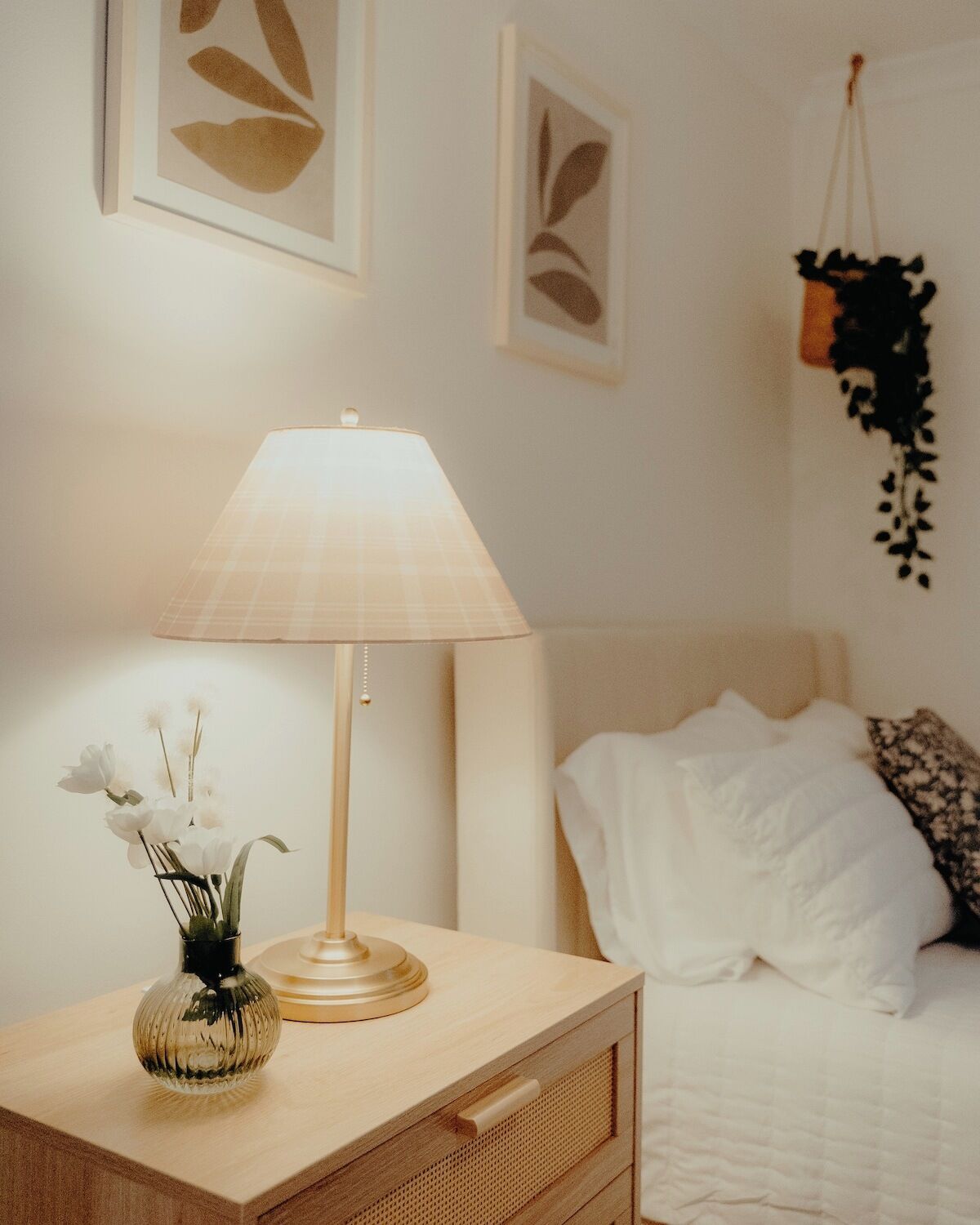 Une chambre à coucher avec une table de chevet, une lampe, un vase de fleurs, un lit avec une literie blanche et des œuvres d’art murales.
