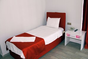 A single bed with a red headboard and white bedding, a bedside table with a phone and a small drawer, and a plain wall with a curtain. - BATIN OTEL (Karabük)