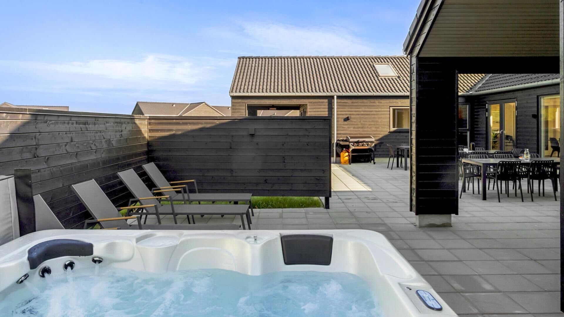 A hot tub with a wooden deck, outdoor seating, and a building with a tiled roof.