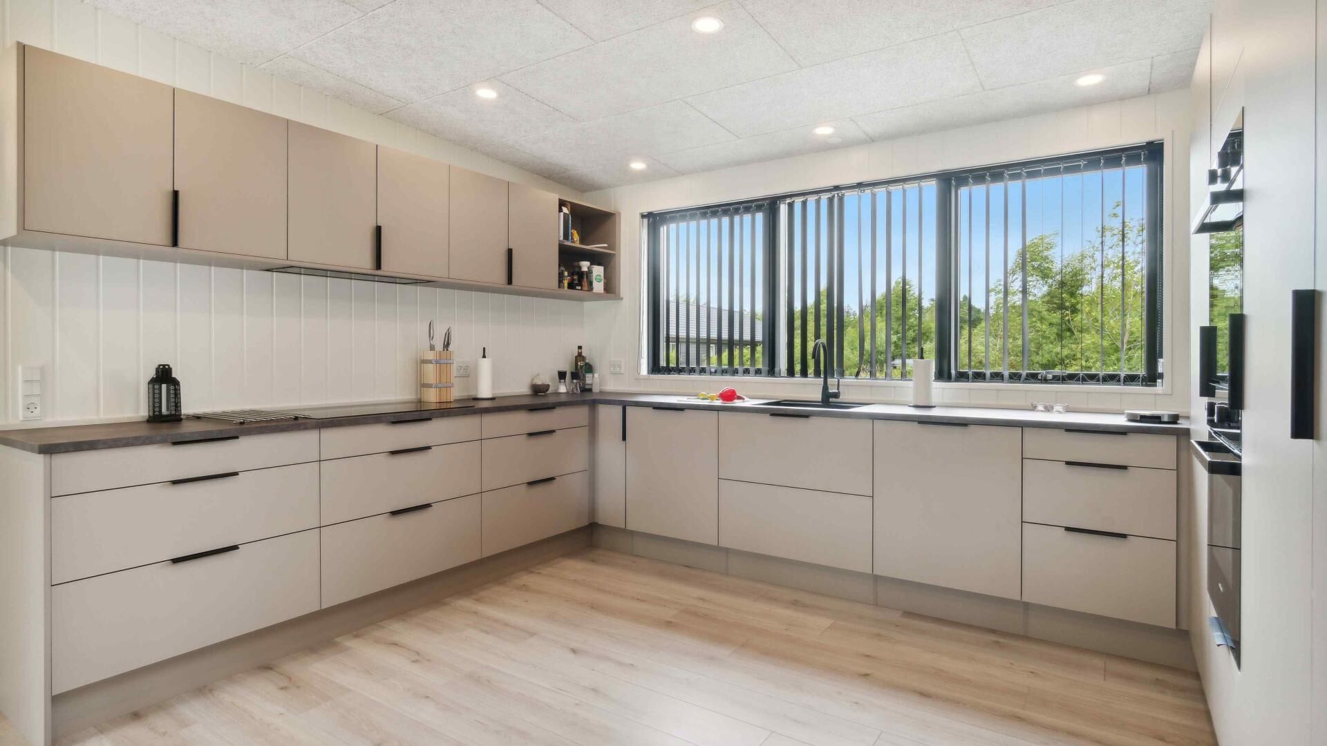 A modern kitchen with light wood cabinetry, a large window with blinds, and a built-in oven.