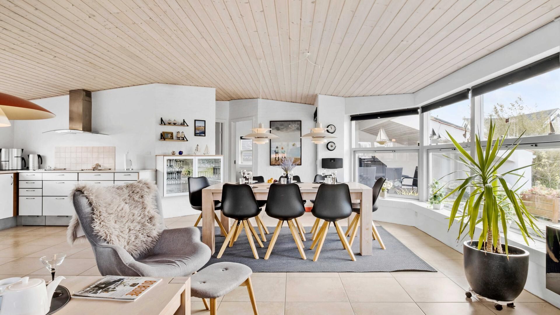 Una cocina moderna con armarios blancos y techo de madera, un comedor con sillas negras y una mesa de madera, y un ventanal con vistas al exterior.