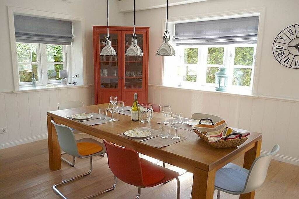 Un coin repas avec une table en bois, des chaises rouges et bleues, des lampes suspendues et un buffet rouge aux portes en verre.