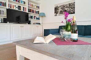 Ein Wohnzimmer mit Fernseher, Bücherregalen, einer Couch, einem Couchtisch mit einem Buch, einer Weinflasche und einer Vase mit Blumen.