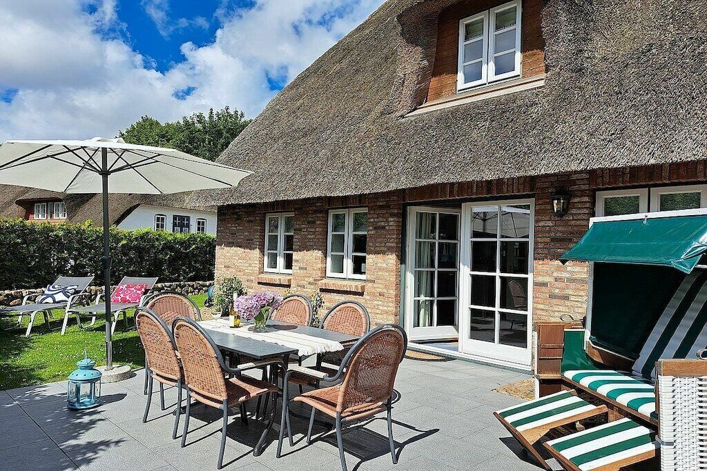 Ein Haus mit Strohdach und einer Terrasse, auf der sich ein Tisch mit Stühlen, eine Liege und ein kleiner Tisch mit einer Laterne befinden.
