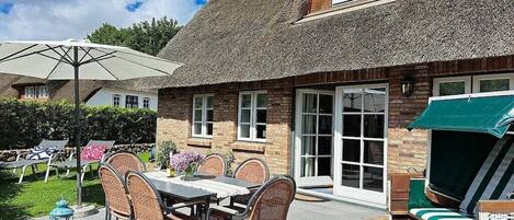 A thatched-roof house with a patio area featuring a table and chairs, a lounge chair, and a small table with a lantern.