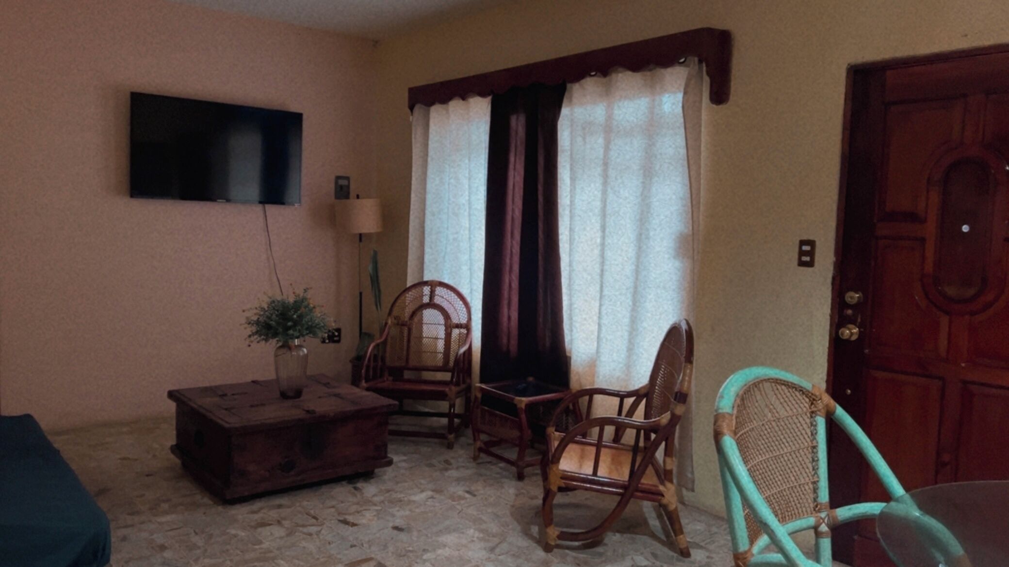 Una habitación con un cofre de madera, dos sillas de madera, un televisor montado en la pared y una puerta.