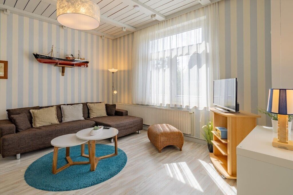 Una sala de estar con papel tapiz a rayas, un sofá marrón, una mesa de centro redonda, un televisor sobre un soporte de madera y un ventanal con cortinas.