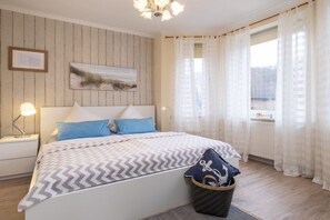 A bedroom with a bed, bedside lamps, a nightstand, a window with curtains, and a wall-mounted painting. - Holiday apartment with 1 bedroom (Travemünde)