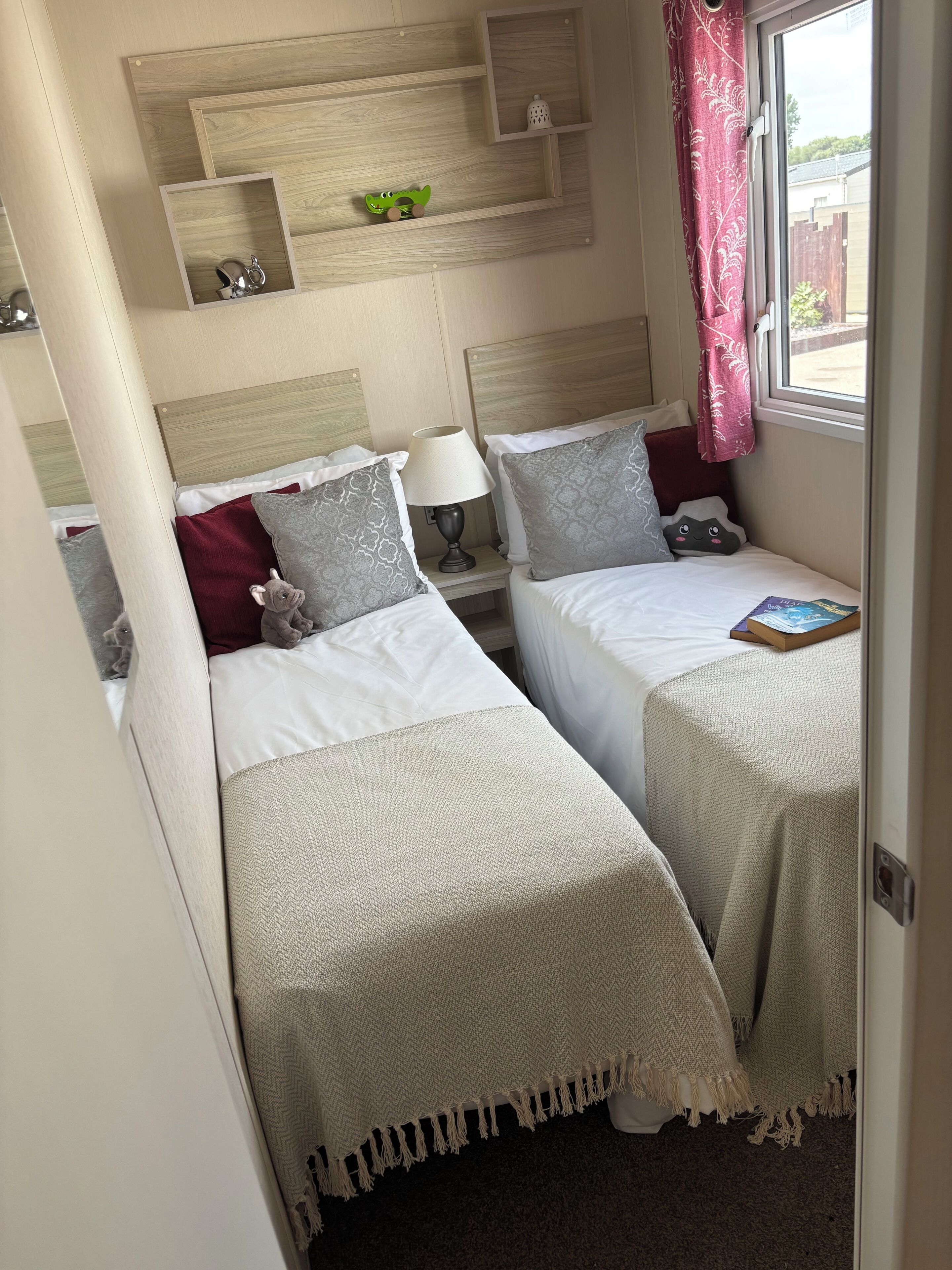 Una habitación de hotel con dos camas, una mesita de noche con lámpara, un estante con objetos decorativos y una ventana con vistas.