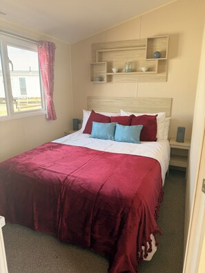 Una habitación de hotel con cama, ventana con cortinas, mesita de noche con lámpara y estante con objetos decorativos.