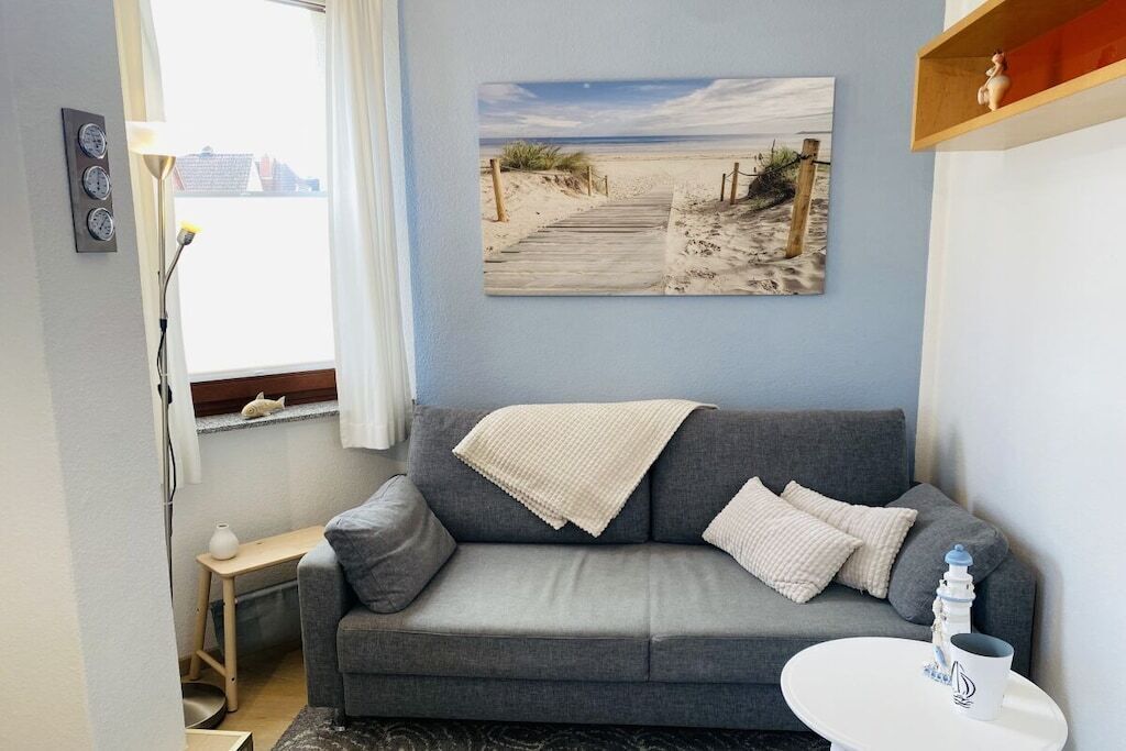 Een woonkamer met een grijze bank, een kleine tafel, een schilderij van een strand en een raam met gordijnen.