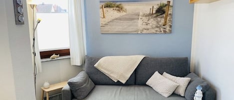 Een woonkamer met een grijze bank, een kleine tafel, een schilderij van een strand en een raam met gordijnen.