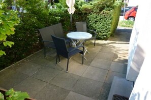 Eine Terrasse im Freien mit einem runden Tisch und zwei Stühlen, umgeben von Grünpflanzen und einem Hecken.